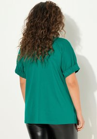 Donna con lunghi capelli ricci che indossa una maglietta verde a maniche corte e larghe e pantaloni in pelle neri, mostrata di spalle su uno sfondo bianco.