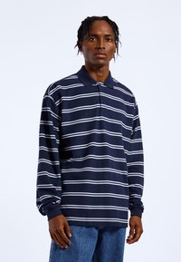 Navy blue long-sleeve polo shirt with white horizontal stripes, featuring a collar and button front, made of textured fabric, paired with denim jeans.