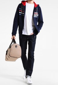 Marinblå zip-hoodie med röda detaljer och randiga inslag, vit t-shirt, mörka jeans och vita sneakers; håller en beige duffelväska med svarta detaljer.