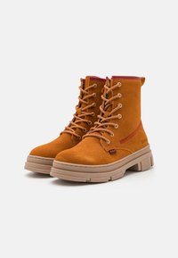 Brown suede ankle boots with a round toe, featuring a beige rubber sole, six eyelet lacing, and red accents on the sides and top.