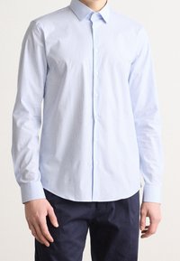 Camisa de botones de manga larga en azul claro con un patrón de rayas sutil, cuello clásico y silueta ajustada. La tela parece suave y nítida.