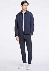 Giacca blu navy con bottoni, dotata di una tasca sul petto, abbinata a pantaloni slim-fit coordinati e sneakers bianche. Texture del tessuto liscia.