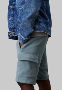 Helle blaue Cargo-Shorts mit Seitentaschen, versehen mit dem "Calvin Klein"-Logo, kombiniert mit einer Jeansjacke. Glatter Baumwollstoff.