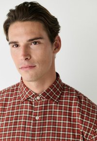 Chemise à carreaux rouge et blanc avec un col classique, la texture du tissu semble douce, présente de petits boutons sur le plastron.
