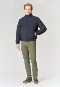 Giacca bomber navy con colletto alto, zip frontale e polsini aderenti. Abbinata a pantaloni verde oliva e scarpe sportive scure.