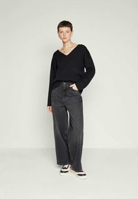 Woman with short hair wearing a black V-neck sweater, wide-leg black jeans, and white sneakers with red and black accents, standing against a plain background.