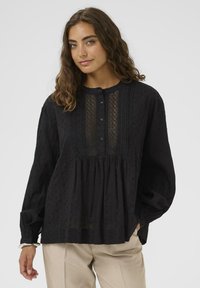 Eine Frau mit lockigem braunem Haar trägt eine lockere schwarze Bluse mit transparentem Vorderteil und beigen Hosen, während sie vor einem schlichten, hellen Hintergrund steht.