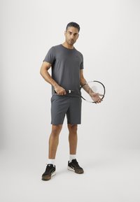 Mann i mørkegrå treningsklær som holder en tennisracket, stående mot en ensfarget hvit bakgrunn.