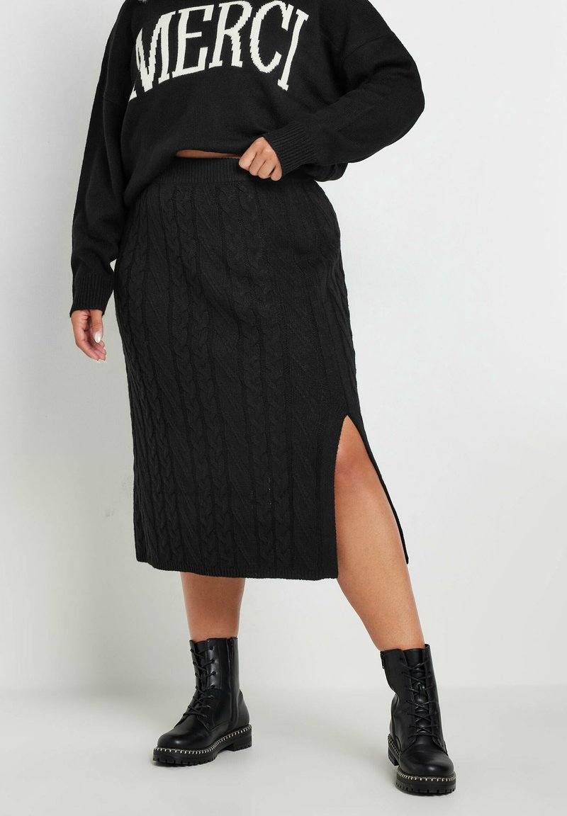 Mustat neulotut midi-hameet, joissa on kaapelikuviot ja sivuaukko, yhdistettynä mustiin nauhalla varustettuihin taistelukenkään ja mustaan neulepaitaan, jossa on valkoista tekstiä.