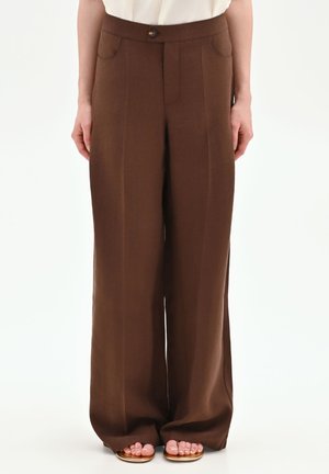 Donna che indossa pantaloni marroni a vita alta e gamba larga con tasche frontali e bottone, abbinati a sandali aperti sulle dita, su sfondo bianco.