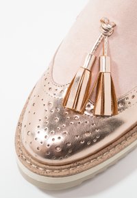 Chaussure métallique en or rose avec un design perforé et deux pompons sur une tige en suède doux, une semelle texturée et une bordure en caoutchouc contrastante.
