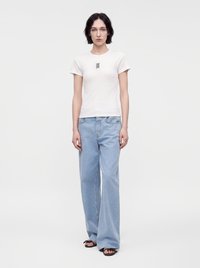 Femme aux cheveux courts et foncés portant un t-shirt blanc à manches courtes, un jean large bleu clair et des sandales noires, debout devant un fond uni.