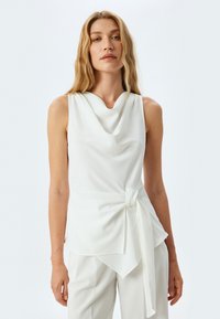 Top bianco senza maniche con scollatura drappeggiata, dettaglio a nodo in vita e tessuto morbido con silhouette fluida.