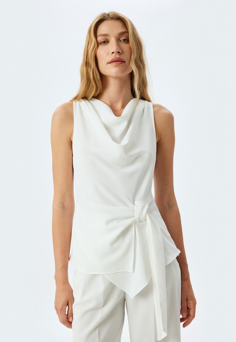 Top bianco senza maniche con scollatura drappeggiata, dettaglio a nodo in vita e tessuto morbido con silhouette fluida.