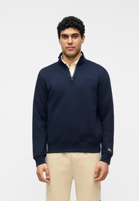 Homme portant un sweat-shirt marine à fermeture éclair courte sur un pantalon beige, debout devant un fond clair uni, faisant face à l'avant avec les bras détendus.