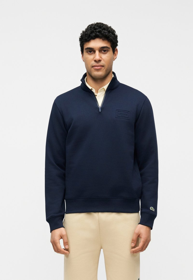 Homme portant un sweat-shirt marine à fermeture éclair courte sur un pantalon beige, debout devant un fond clair uni, faisant face à l'avant avec les bras détendus.