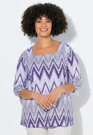 Nainen, jolla on kiharat hiukset, pukeutuneena violetti-valkoiseen siksak-kuvioiseen yläosaan, jossa on kolme neljänneksen hihaa, hymyilee ja pitää käsiään edessään.