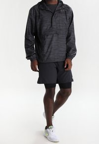 Mężczyzna w czarnej kurtce typu windbreaker w kamuflażu, czarnych warstwowych szortach i białych trampkach Nike stoi na białym tle.