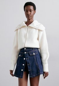 White zip-up sweater with large collar adorned with decorative embellishments, paired with a dark denim skirt featuring front buttons and pockets.