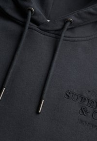 Sudadera con capucha gris oscuro con cordones gruesos que terminan en puntas de metal, con un texto bordado sutilmente "Superdry & Co New York" en el pecho.