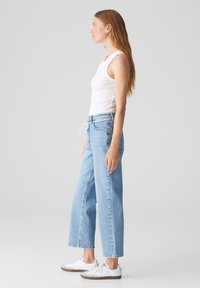 Vrouw in een witte tanktop en lichtblauwe wijde spijkerbroek. De spijkerbroek heeft een trekkoord in de taille en is gecombineerd met witte sneakers.