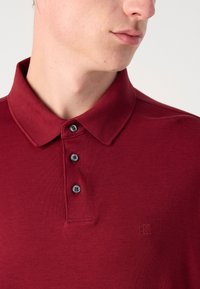 Burgundy színű póló, lélegző anyagból készült, klasszikus gallérral, három sötét gombbal, és egy diszkrét CK logóval a mellrészen.