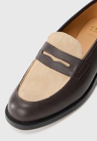 Herrens loafers med mörkbrunt läder och beige mockaöverdel som har en dekorativ läderrem över toppen.