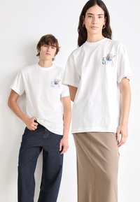 Hvide bomulls-T-shirts med blomsterprint på venstre bryst. Pasformen er afslappet; en model har blå jeans på, den anden har en brun nederdel på.