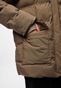 Chaqueta acolchada marrón con una mano visible metiéndose en un bolsillo frontal de parche. La tela parece lisa y ligeramente brillante.