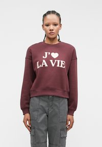 Burgundy sweatshirt with white text "J'♥ LA VIE," ribbed neckline, and cuffs. Paired with grey cargo pants featuring multiple pockets.