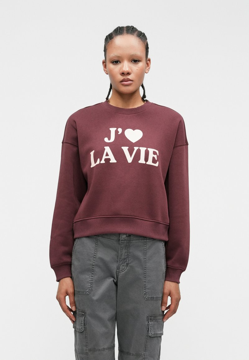 Burgundy sweatshirt with white text "J'♥ LA VIE," ribbed neckline, and cuffs. Paired with grey cargo pants featuring multiple pockets.