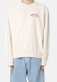 Crème-kleurige sweatshirt met een ronde hals, geribbelde mouwen en onderkant, voorzien van rode geborduurde tekst op de voorkant, gecombineerd met lichtblauwe jeans.