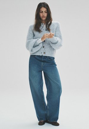 Gaiši zila, teksturēta adīta jaka ar piecām pogām. Kombinēta ar vaļīgām tumši zilām džinsa biksēm. Modele pielāgo savu plaukstas locītavu. Vienkāršs fonu.