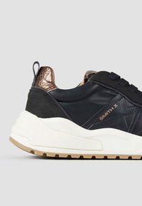 Schwarze Leder- und Wildlederschuhe mit metallisch bronze Akzenten, dicke weiße Sohle, strukturierte Details und flachen Schnürsenkeln.