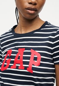 Personne portant un t-shirt rayé bleu marine et blanc avec de grandes lettres rouges « GAP » sur la poitrine, montrant le cou et le bas du visage.