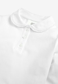 Vit polo-tröja med en volangkrage och en knapp-up placket med tre knappar. Tillverkad av mjukt tyg med en slät yta.
