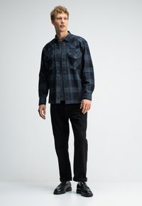 Zwart geruit shirt met drukknoopjes, voorzien van twee borstzakken, gedragen over een grijze T-shirt, in combinatie met zwarte broek en zwarte schoenen.