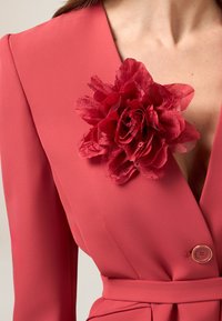 Korallenfarbener Blazer mit tiefem V-Ausschnitt, langen Ärmeln und einem strukturierten Stoff. Verzückt mit einer großen roten Stoffblumen-Brosche.