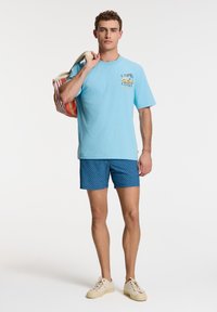 Svijetloplavi T-shirt s grafičkim printom, uparen s tamnoplavim kratkim hlačama sa uzorkom. Bež cipele i torba sa višebojnim prugama.