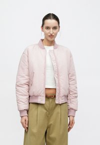 Giacca bomber imbottita in rosa tenue con polsini e orlo a coste, dotata di cerniera frontale e tasche laterali. Indossata sopra un crop top bianco.