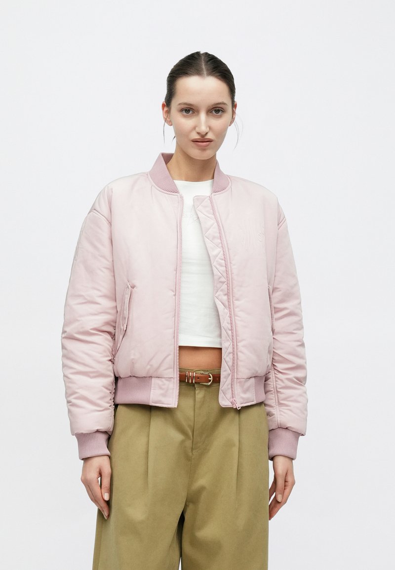 Giacca bomber imbottita in rosa tenue con polsini e orlo a coste, dotata di cerniera frontale e tasche laterali. Indossata sopra un crop top bianco.