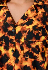 Camicia a maniche corte con motivo a fiamma nera e arancione, dotata di taschino sul petto e bottoni neri. Tessuto morbido e liscio con un design vivace.