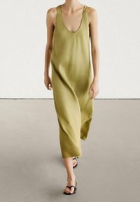 Olivegrünes ärmelloses Kleid mit tiefem V-Ausschnitt, fließender Form und weicher Textur, das bis zur Mitte der Waden reicht. Kombiniert mit zarten Sandalen.