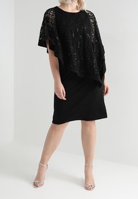 Frau in einem schwarzen knielangen Kleid mit einem transparenten Spitzenüberzug aus Pailletten und silbernen High-Heels, die vor einem weißen Hintergrund steht.