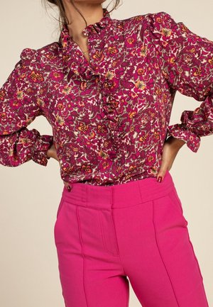 Kvinde iført en langærmet bluse med blomsterprint og flæser, kombineret med højtaljede, knaldrøde bukser, hænderne på hofterne, foran en ensfarvet baggrund.