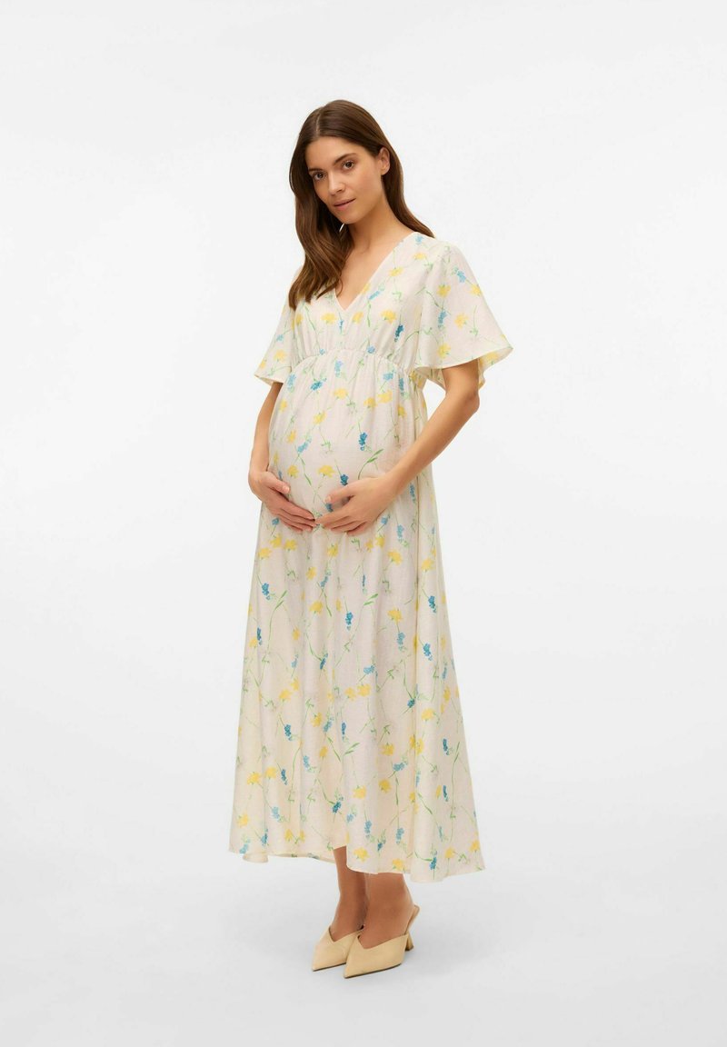 Robe de maternité en tissu crème avec un motif floral jaune et bleu, décolleté en V, manches courtes flottantes et taille froncée. Portée avec des talons beiges.
