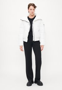 Witte pufferjack met een capuchon, ritssluiting en elastische manchet. Draag het over een zwarte top en combineer het met zwarte, ruime broek en zware zwarte schoenen.