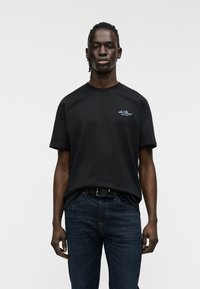 Μαύρο βαμβακερό μπλουζάκι με στρογγυλή λαιμόκοψη, κοντά μανίκια και μικρό λευκό λογότυπο στο αριστερό στήθος. Φορεμένο με σκούρο μπλε τζιν παντελόνι.