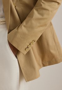 Μανίκι μπεζ σακακιού με τέσσερα κουμπιά, φορεμένο πάνω από λευκό σύνολο, με το χέρι μερικώς στην τσέπη, που δείχνει την υφή του υφάσματος και τις ραφές.