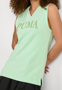 Kvinna som bär en ärmlös mintgrön Puma-topp med V-ringning och svarta byxor, visar arm-tatueringar och har lockigt hår.